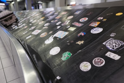 A luggage conveyor belt is covered with various colorful stickers. The surface is metallic and black, providing contrast to the stickers. Each sticker has unique designs and logos, and a piece of luggage is also partially visible on the conveyor.