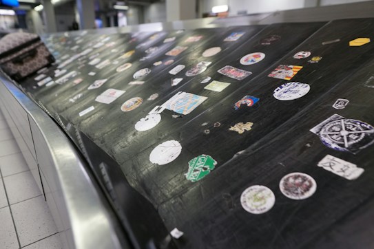 A luggage conveyor belt is covered with various colorful stickers. The surface is metallic and black, providing contrast to the stickers. Each sticker has unique designs and logos, and a piece of luggage is also partially visible on the conveyor.