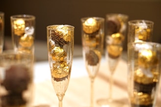 Several champagne glasses are filled with golden wrapped chocolates. The composition is focused on the elegant display of the chocolates, with their shiny golden wrappers creating a luxurious and festive atmosphere. The background appears to be blurred, drawing attention to the glasses.