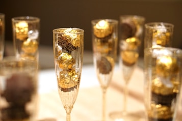 Several champagne glasses are filled with golden wrapped chocolates. The composition is focused on the elegant display of the chocolates, with their shiny golden wrappers creating a luxurious and festive atmosphere. The background appears to be blurred, drawing attention to the glasses.