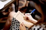 A person is holding a pen and writing on a printed sheet titled 'He said She said' with various questions or prompts written on it. The sheet is resting on the person's lap, and they are wearing a patterned black and white skirt. The pen is blue, and the scene appears to be in an indoor setting with a carpeted floor.