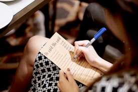 A person is holding a pen and writing on a printed sheet titled 'He said She said' with various questions or prompts written on it. The sheet is resting on the person's lap, and they are wearing a patterned black and white skirt. The pen is blue, and the scene appears to be in an indoor setting with a carpeted floor.