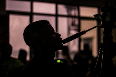 A silhouette of a person speaking into a microphone with a camera in the foreground. The background is blurred with several figures and a large window.