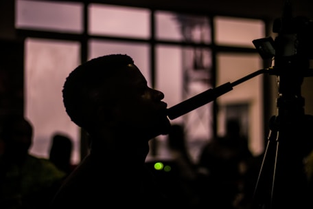 A silhouette of a person speaking into a microphone with a camera in the foreground. The background is blurred with several figures and a large window.
