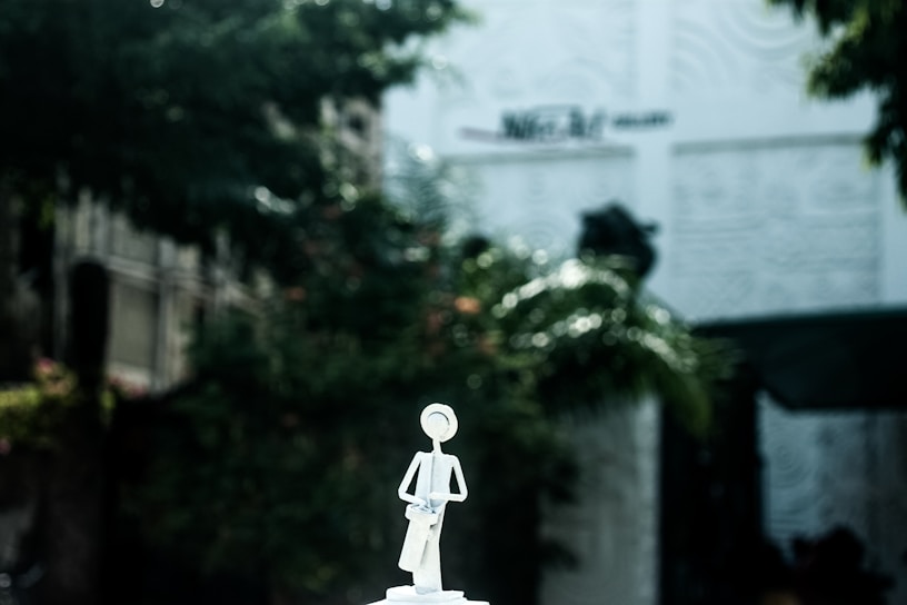 A minimalist sculpture of a figure is positioned against a blurry background of greenery and architectural elements. The figure appears to be stylized and abstract, possibly made from metal or similar material, standing on a pedestal.
