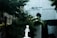 A minimalist sculpture of a figure is positioned against a blurry background of greenery and architectural elements. The figure appears to be stylized and abstract, possibly made from metal or similar material, standing on a pedestal.