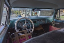 The interior of a vintage car with an old-style steering wheel wrapped in a cover sits in front of a retro dashboard with analog gauges. The view through the windshield shows a street scene with a woman walking and a parked blue motorcycle. A decorative hanging ornament dangles from the rearview mirror.