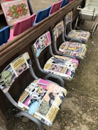 A row of chairs with retro comic book upholstery featuring colorful and vintage comic strip designs. These chairs are placed against a wooden barrier. In the background, there are vibrant blue and pink seats, as well as an ornate decorative panel. A vintage white cart is visible to one side.