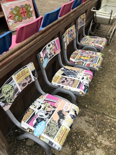 A row of chairs with retro comic book upholstery featuring colorful and vintage comic strip designs. These chairs are placed against a wooden barrier. In the background, there are vibrant blue and pink seats, as well as an ornate decorative panel. A vintage white cart is visible to one side.