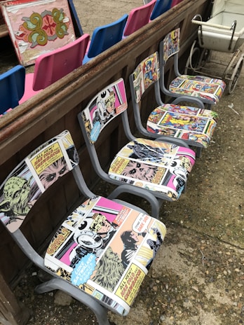 A row of chairs with retro comic book upholstery featuring colorful and vintage comic strip designs. These chairs are placed against a wooden barrier. In the background, there are vibrant blue and pink seats, as well as an ornate decorative panel. A vintage white cart is visible to one side.