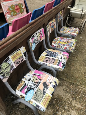 A row of chairs with retro comic book upholstery featuring colorful and vintage comic strip designs. These chairs are placed against a wooden barrier. In the background, there are vibrant blue and pink seats, as well as an ornate decorative panel. A vintage white cart is visible to one side.
