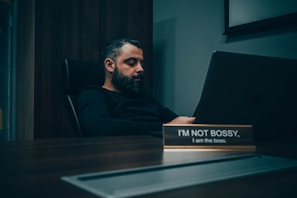 A man with a beard is sitting at a desk in a dimly lit office, looking at a laptop. The desk has a nameplate that reads 'I'M NOT BOSSY, I am the boss.' The atmosphere is calm and introspective.
