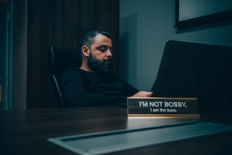 A man with a beard is sitting at a desk in a dimly lit office, looking at a laptop. The desk has a nameplate that reads 'I'M NOT BOSSY, I am the boss.' The atmosphere is calm and introspective.