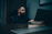 A man with a beard is sitting at a desk in a dimly lit office, looking at a laptop. The desk has a nameplate that reads 'I'M NOT BOSSY, I am the boss.' The atmosphere is calm and introspective.
