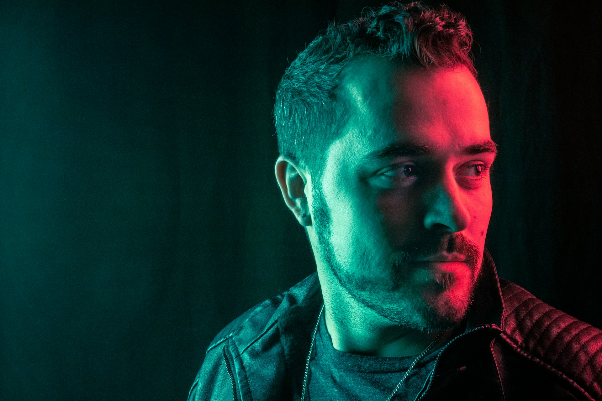 A man with short hair and a beard is seen in a dramatic lighting setup, featuring a stark contrast between red and teal lighting on his face. He is wearing a dark leather jacket, looking off to the side with a thoughtful expression.