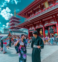 People in traditional Japanese attire are gathered in a bustling temple area with vibrant red architecture. The scene is lively, filled with tourists and locals exploring the historic site, against a backdrop of a pagoda and a bright blue sky dotted with clouds.