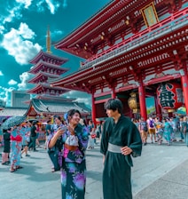 People in traditional Japanese attire are gathered in a bustling temple area with vibrant red architecture. The scene is lively, filled with tourists and locals exploring the historic site, against a backdrop of a pagoda and a bright blue sky dotted with clouds.