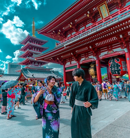 People in traditional Japanese attire are gathered in a bustling temple area with vibrant red architecture. The scene is lively, filled with tourists and locals exploring the historic site, against a backdrop of a pagoda and a bright blue sky dotted with clouds.