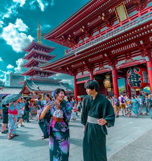 People in traditional Japanese attire are gathered in a bustling temple area with vibrant red architecture. The scene is lively, filled with tourists and locals exploring the historic site, against a backdrop of a pagoda and a bright blue sky dotted with clouds.