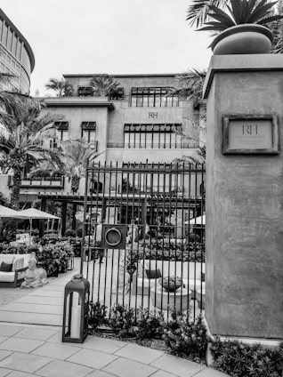 An upscale building surrounded by palm trees and intricate metal gates. The front yard is decorated with outdoor furniture, potted plants, and a large lantern. The architecture features large windows and a modern aesthetic.
