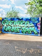A vibrant street art mural with large, bold graffiti letters spelling 'Grenfell Graff Jam' in green and white against a blue wall. Surrounding the text are artistic elements such as stars and a sky-like pattern. The scene is set outdoors with trees and a partly cloudy sky in the background, and the ground is paved with bricks.