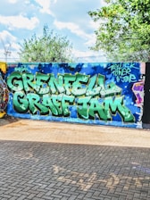 A vibrant street art mural with large, bold graffiti letters spelling 'Grenfell Graff Jam' in green and white against a blue wall. Surrounding the text are artistic elements such as stars and a sky-like pattern. The scene is set outdoors with trees and a partly cloudy sky in the background, and the ground is paved with bricks.