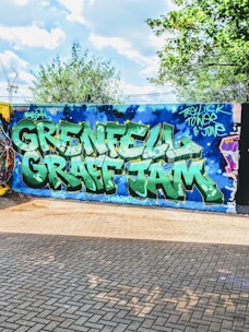 A vibrant street art mural with large, bold graffiti letters spelling 'Grenfell Graff Jam' in green and white against a blue wall. Surrounding the text are artistic elements such as stars and a sky-like pattern. The scene is set outdoors with trees and a partly cloudy sky in the background, and the ground is paved with bricks.