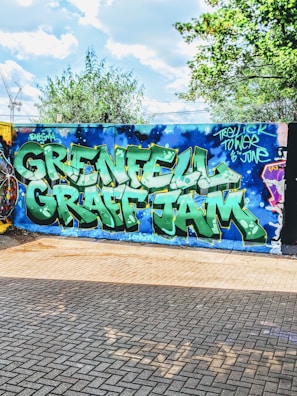 A vibrant street art mural with large, bold graffiti letters spelling 'Grenfell Graff Jam' in green and white against a blue wall. Surrounding the text are artistic elements such as stars and a sky-like pattern. The scene is set outdoors with trees and a partly cloudy sky in the background, and the ground is paved with bricks.