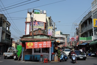A busy street scene in an urban area features a small food vendor stall with a prominent red sign displaying menu items and prices in Chinese characters. Surrounding the stall are motorcycles, a truck, and several buildings adorned with various colorful signs. Numerous cables crisscross the sky above, and people can be seen riding scooters and walking in the area.