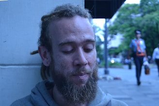 A person with a beard and dreadlocks is leaning against a light-colored wall. Their eyes are closed, and they have a pensive expression. The background shows an outdoor scene with trees, a person walking, and buildings in the distance.