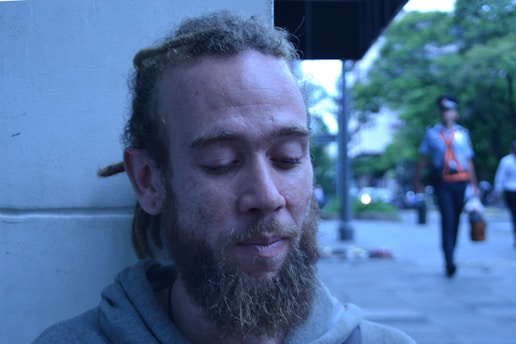 A person with a beard and dreadlocks is leaning against a light-colored wall. Their eyes are closed, and they have a pensive expression. The background shows an outdoor scene with trees, a person walking, and buildings in the distance.