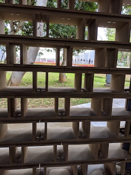 A wooden partition with rectangular cutouts allows a view of the outdoor scenery beyond. Trees and greenery are visible through the openings, with a hint of flowers and distant structures.