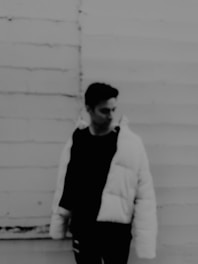 Still frame from a grainy black and white film showing a solitary figure against a textured wall.