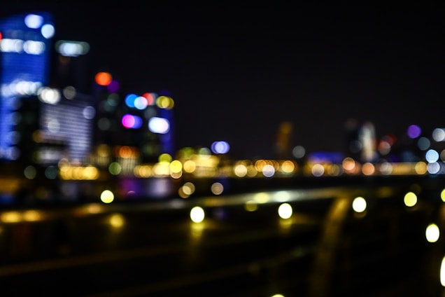 A blurry cityscape at night with multicolored lights and outlines of skyscrapers. The scene is out of focus, lending a dreamy and abstract quality to the urban setting.