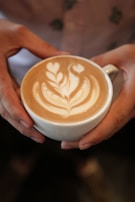 A close-up of hands gently holding a warm latte with intricate leaf art on top.