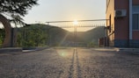 A gate made of metal bars is positioned in front of a sunrise or sunset, with the sun casting long shadows on a paved surface. To the right, there is a building with an AC unit affixed on the exterior wall. Trees and hills are visible in the background.