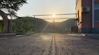 A gate made of metal bars is positioned in front of a sunrise or sunset, with the sun casting long shadows on a paved surface. To the right, there is a building with an AC unit affixed on the exterior wall. Trees and hills are visible in the background.