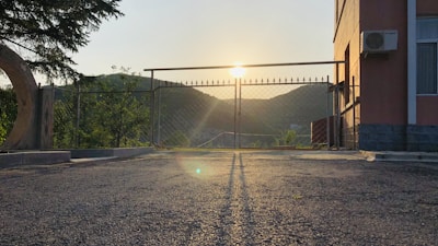 A gate made of metal bars is positioned in front of a sunrise or sunset, with the sun casting long shadows on a paved surface. To the right, there is a building with an AC unit affixed on the exterior wall. Trees and hills are visible in the background.