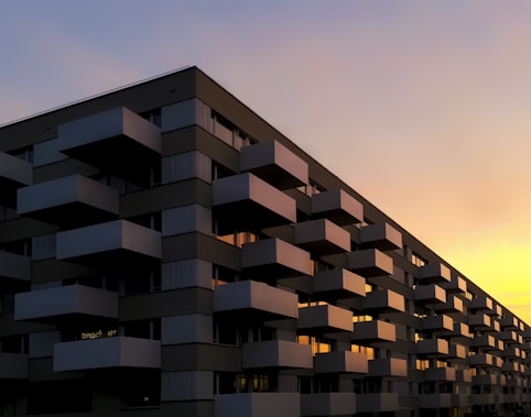 A modern apartment building with a geometric design of protruding balconies against a sunset sky. The structure features a repetitive pattern, creating a sense of depth and symmetry.
