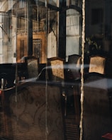 Sunlit interior of a room visible through a glass window, showing several ornate chairs with patterned upholstery. The reflection on the glass creates a layered effect with exterior elements like a door and building features.