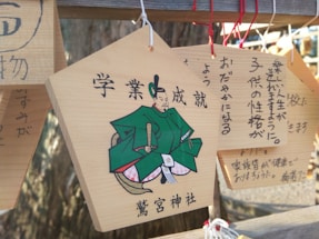 Wooden ema plaques are hanging, featuring Japanese characters and illustrations. One plaque prominently displays a drawing of a figure in traditional Japanese attire, with text written in Japanese around it.