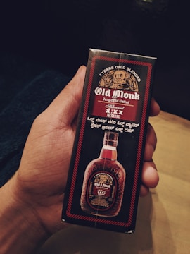 A hand is holding a rectangular box packaging of Old Monk rum, featuring the brand name in bold white letters on a black background. The design includes a circular emblem of a monk's face and additional text in a smaller font. Below the text, there is an image of the bottle itself, showcasing its distinctive shape.