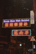 Neon signs illuminate a city street with bright colors and text, set against the backdrop of a dark, towering building with lit windows. The signs display both English and Chinese characters, indicating a bridal shop. A small pedestrian sign is visible in the foreground.