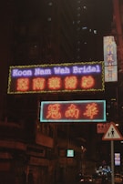 Neon signs illuminate a city street with bright colors and text, set against the backdrop of a dark, towering building with lit windows. The signs display both English and Chinese characters, indicating a bridal shop. A small pedestrian sign is visible in the foreground.