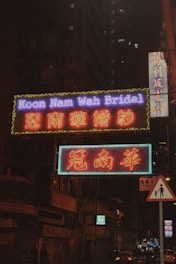 Neon signs illuminate a city street with bright colors and text, set against the backdrop of a dark, towering building with lit windows. The signs display both English and Chinese characters, indicating a bridal shop. A small pedestrian sign is visible in the foreground.