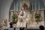 A religious ceremony is taking place with a priest in ornate vestments raising the Eucharist in front of a beautifully decorated altar. The altar is adorned with white flowers and tall candle holders. A statue of an angel is visible on the side, and several lit candles are placed around.