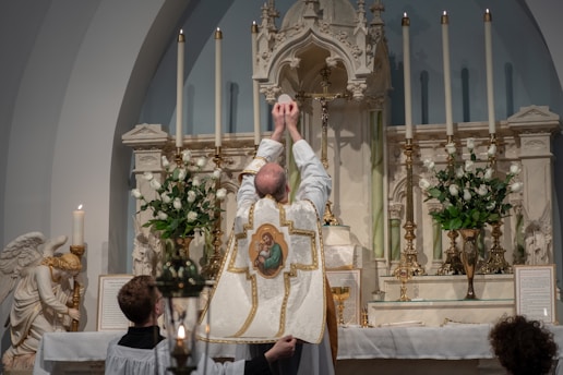 A religious ceremony is taking place with a priest in ornate vestments raising the Eucharist in front of a beautifully decorated altar. The altar is adorned with white flowers and tall candle holders. A statue of an angel is visible on the side, and several lit candles are placed around.