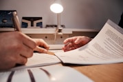 Two hands are visible, one holding a pen and another holding a document, suggesting a situation involving document review or signing. The scene appears to be set on a desk with a table lamp illuminating the area.