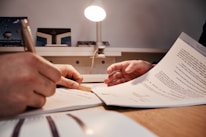 Two hands are visible, one holding a pen and another holding a document, suggesting a situation involving document review or signing. The scene appears to be set on a desk with a table lamp illuminating the area.