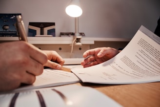 Two hands are visible, one holding a pen and another holding a document, suggesting a situation involving document review or signing. The scene appears to be set on a desk with a table lamp illuminating the area.